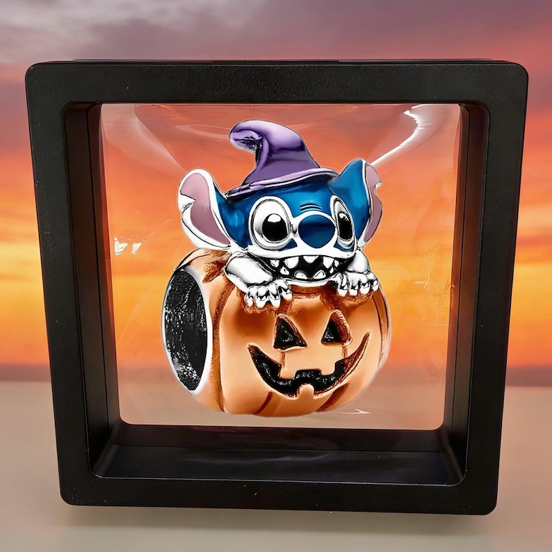 May include: A metal charm bead featuring a blue character with large ears wearing a purple pointed hat, resting on top of an orange jack-o'-lantern pumpkin. The charm is suspended within a square black floating display frame with a clear film.