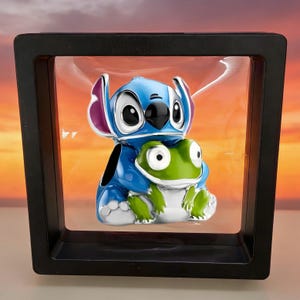 May include: A blue and white enamel charm featuring a cartoon character with large ears holding a small green frog. The charm is suspended within a square black floating display frame that creates a clear view of the character design.