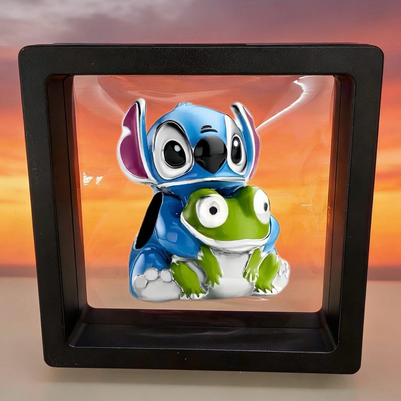 May include: A blue and white enamel charm featuring a cartoon character with large ears holding a small green frog. The charm is suspended within a square black floating display frame that creates a clear view of the character design.