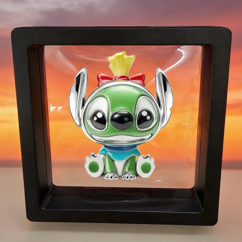 May include: A metallic charm featuring a green character with large black eyes, long ears, and a red bow on its head, wearing a blue collar. The charm is suspended inside a square black floating display frame with a clear plastic membrane.