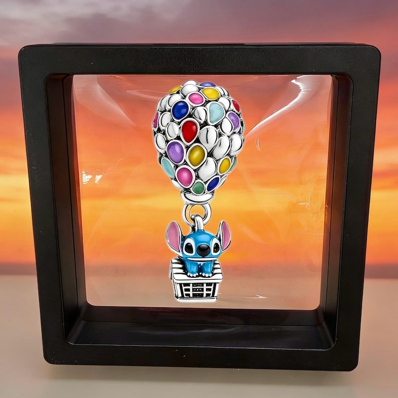 May include: A silver charm featuring a cluster of colorful enamel balloons in red, blue, yellow, purple, and pink, attached to a small blue character with large ears sitting on top of a white and black house structure, suspended in a black frame.