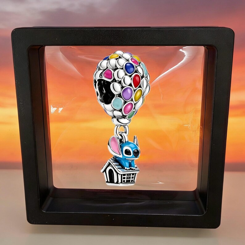 May include: A silver charm featuring a cluster of colorful enamel balloons in red, yellow, blue, purple, and teal, suspended above a blue character figure sitting on top of a small silver doghouse with black detailing and a metallic finish.