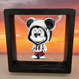 May include: A silver-toned charm featuring a cartoon mouse character wearing a white hooded sweatshirt with black ears and dark red shorts, suspended within a square black floating display frame against a sunset-colored background.