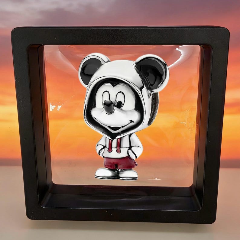 May include: A silver-toned charm featuring a cartoon mouse character wearing a white hooded sweatshirt with black ears and dark red shorts, suspended within a square black floating display frame against a sunset-colored background.