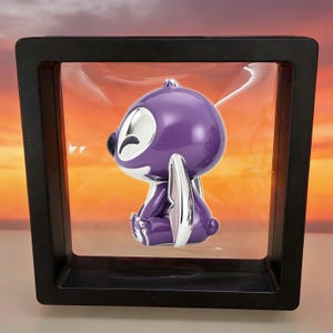 May include: A purple and silver stylized creature figurine suspended inside a square black floating display frame. The figurine features a smooth, glossy finish with large ears and a rounded head, held in place by clear plastic membranes.