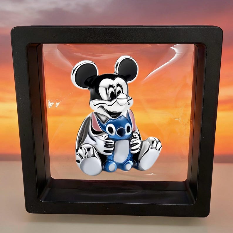 May include: A silver-toned metal charm featuring a black and white Mickey Mouse figure holding a small blue Stitch character. The charm is suspended within a square black floating display frame with a clear, flexible plastic membrane.