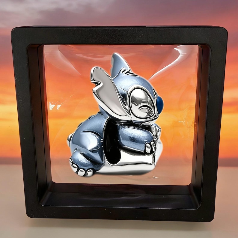 May include: A metallic blue and silver charm shaped like the character Stitch, featuring large ears, a rounded nose, and a smooth, polished finish. The charm is suspended within a square black floating display frame with clear plastic membranes.