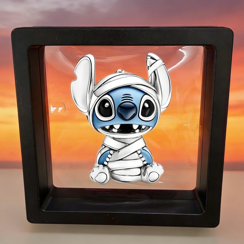 May include: A silver-toned charm featuring the character Stitch wrapped in white bandages. The figure has large ears, a blue face, and black eyes. It is displayed inside a square black floating frame with a clear centre, showcasing the character.