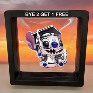 Puede incluir: Un charm plateado con un personaje de dibujos animados con birrete y pergamino. El charm tiene detalles azules y morados. Se muestra en un marco negro con el texto "BYE 2 GET 1 FREE" en la parte superior.