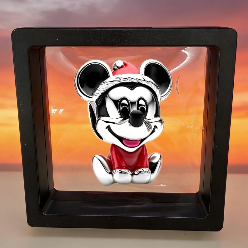May include: A silver-toned Mickey Mouse figurine wearing a red and white Santa hat and a red outfit, suspended within a square black floating display frame. The character has black ears, a smiling expression, and a polished, reflective metallic finish.
