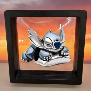 May include: A silver-toned metal charm featuring the character Stitch from Lilo & Stitch, depicted in a sleeping pose with large ears and a blue nose, suspended within a square black floating display frame with a clear plastic membrane.