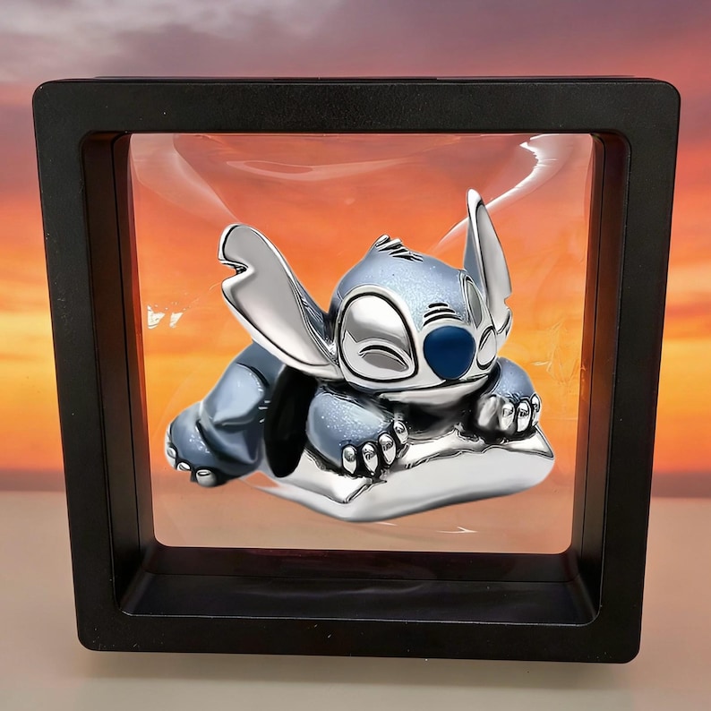May include: A silver-toned metal charm featuring the character Stitch from Lilo & Stitch, depicted in a sleeping pose with large ears and a blue nose, suspended within a square black floating display frame with a clear plastic membrane.