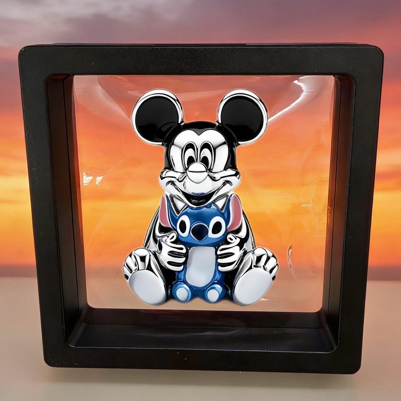 May include: A metallic silver and black figurine of Mickey Mouse holding a small blue and light blue Stitch character, encased within a square black floating display frame with a clear membrane that suspends the object in the center.