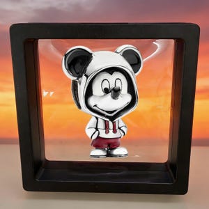 May include: A silver-toned charm featuring a cartoon mouse character wearing a white hoodie with black ears and dark red shorts, suspended within a square black floating display frame against a sunset-colored background.