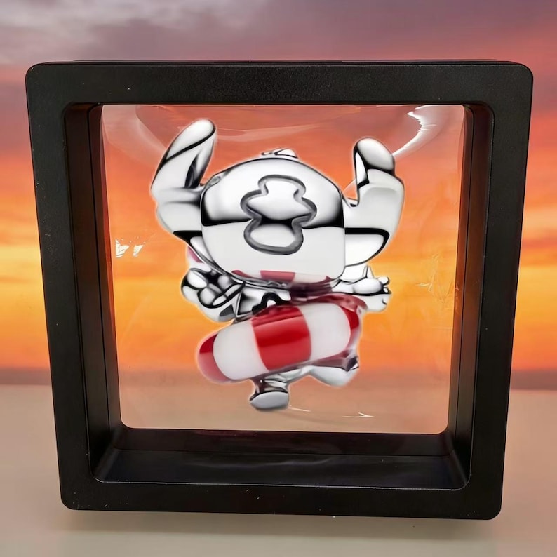 May include: A silver-colored figurine of a cartoon character with large ears and a red and white striped floatation device. The figurine is displayed inside a black shadow box with a clear front, set against a sunset background.