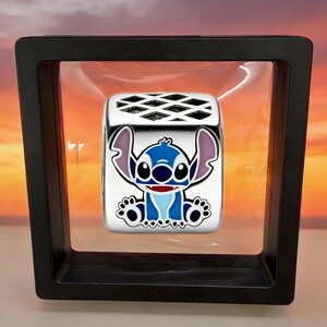 May include: A silver-toned metal charm bead featuring a colorful enamel graphic of the character Stitch. The bead has a square shape with a lattice-patterned top and is suspended within a black square floating display frame with clear film.