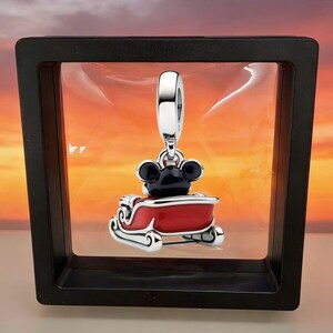 May include: A silver charm featuring a black-eared character sitting in a bright red sleigh with white trim. The charm has a polished silver bail at the top and is suspended within a black square frame against a sunset-colored background.