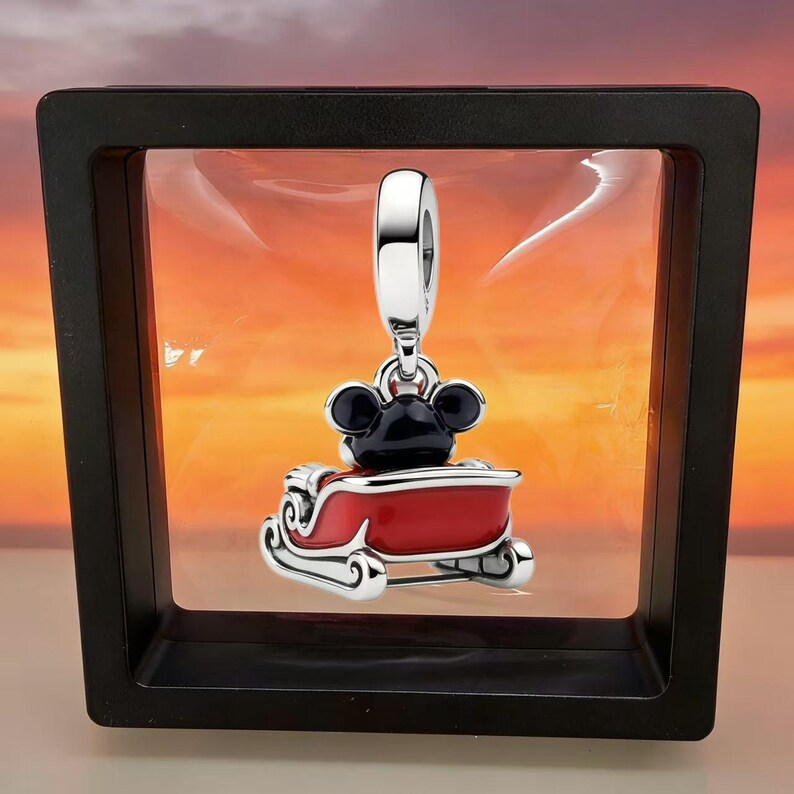 May include: A silver charm featuring a black-eared character sitting in a bright red sleigh with white trim. The charm has a polished silver bail at the top and is suspended within a black square frame against a sunset-colored background.