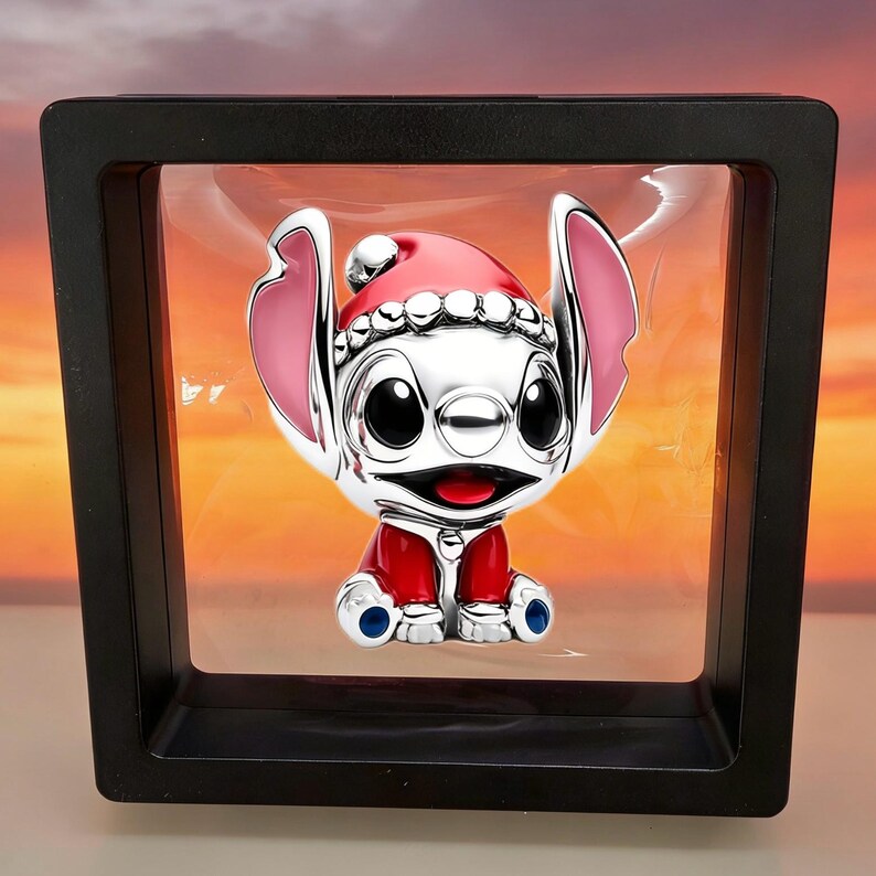 Op de afbeelding: Een zilverkleurig karakterfiguurtje met een rood-witte kerstmuts en een rode outfit, opgehangen in een vierkant zwart zwevend displayframe. Het figuurtje heeft grote zwarte ogen, roze oren en een brede glimlach met open mond.