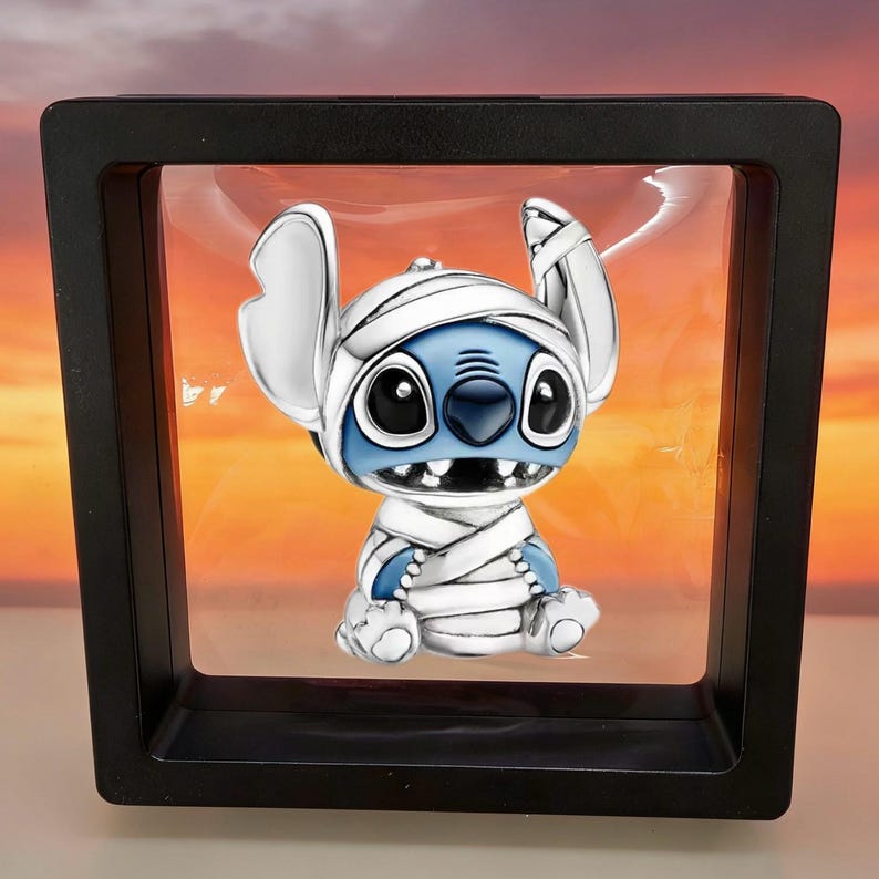 May include: A silver-toned charm featuring the character Stitch wrapped in white bandages like a mummy. The charm has blue enamel details on the face and ears, large black eyes, and a small nose. It is displayed inside a square black frame.