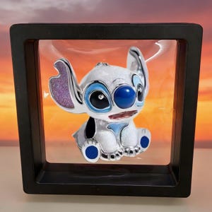 May include: A white, glittery, stylized character figurine with large ears, a blue nose, and black eyes, suspended inside a square black floating display frame. The character features purple inner ear details and blue accents on the feet and eyes.
