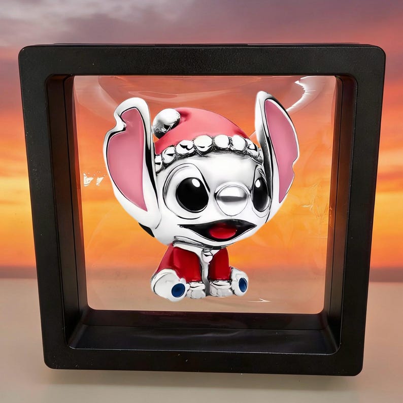 Op de afbeelding: Een zilverkleurige Stitch-bedel met een rood-witte kerstmuts en een rode jas, opgehangen in een vierkant zwart zwevend displayframe. Het personage heeft grote roze oren, zwarte ogen en een wijd open mond met een rode tong zichtbaar.