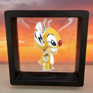 May include: A yellow and silver metallic character figurine with large ears and a red nose, suspended inside a square black floating display frame. The character has a smooth, glossy finish and is positioned in the center of the transparent display.