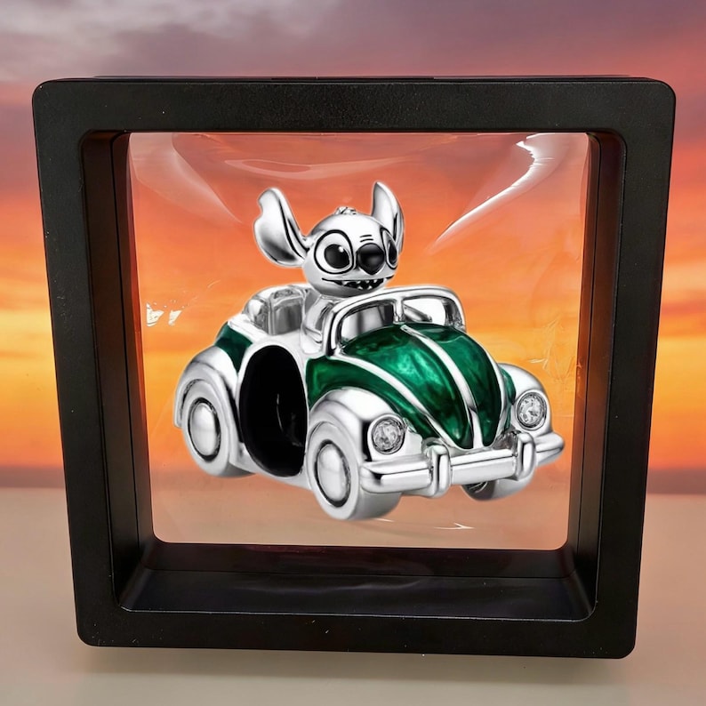 Op de afbeelding: Een zilverkleurige bedel in de vorm van een klassieke auto met een groene emaille afwerking, met het personage Stitch op de bestuurdersstoel. De bedel hangt in een vierkante zwarte zwevende displaylijst met doorzichtig plastic folie.