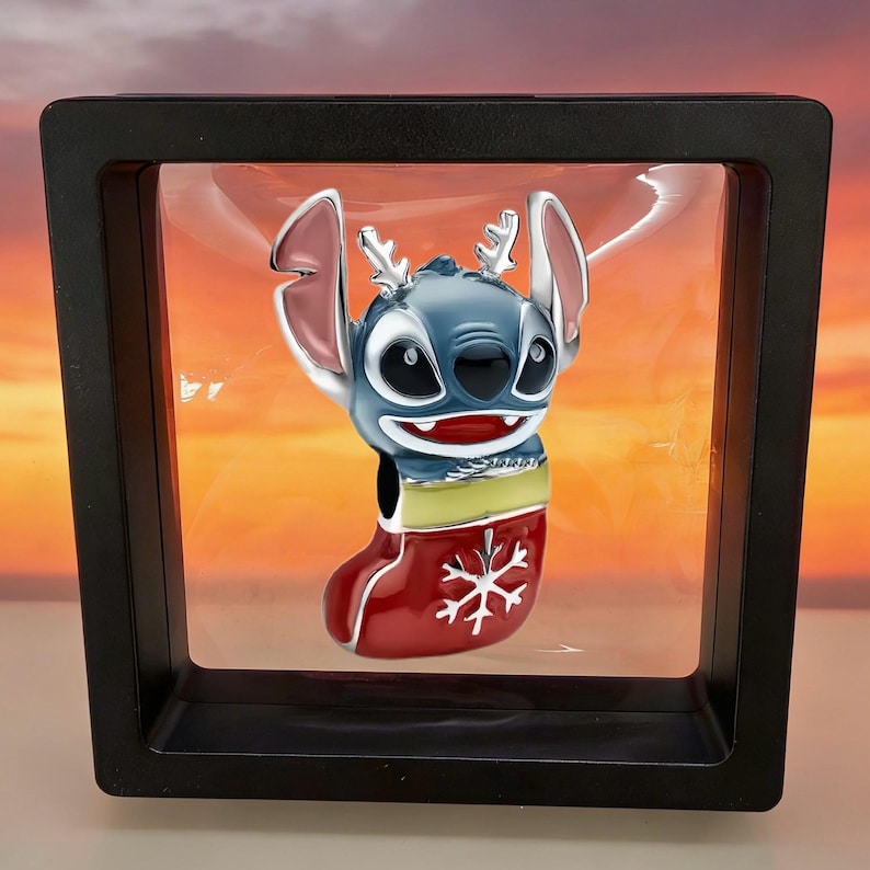 May include: A silver-toned charm featuring a blue character with large ears and reindeer antlers, emerging from a red Christmas stocking decorated with a white snowflake pattern, suspended within a square black floating display frame.