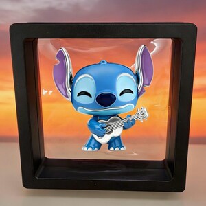 Puede incluir: Una figura de personaje azul con orejas grandes y expresi&oacute;n sonriente, sosteniendo una peque&ntilde;a guitarra blanca. La figura est&aacute; suspendida dentro de un marco de exhibici&oacute;n negro cuadrado con una membrana de pl&aacute;stico transparente.
