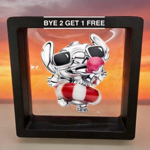 May include: A silver-colored charm in a black display case. The charm depicts a cartoon character wearing sunglasses and holding a pink ice cream cone. The character is also wearing a red and white striped floatation device. The text "BYE 2 GET 1 FREE" is at the top.