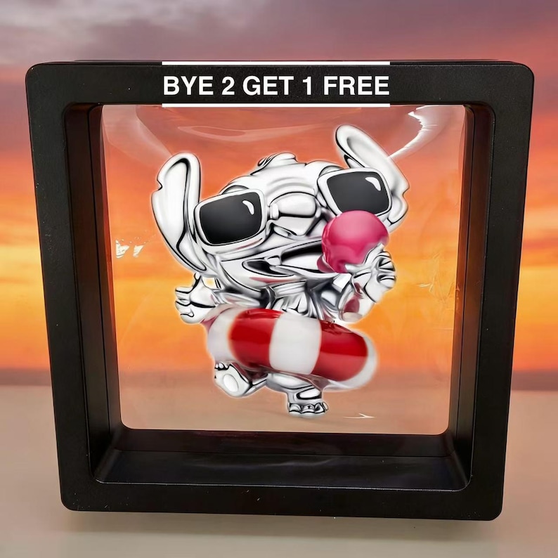 May include: A silver-colored charm in a black display case. The charm depicts a cartoon character wearing sunglasses and holding a pink ice cream cone. The character is also wearing a red and white striped floatation device. The text "BYE 2 GET 1 FREE" is at the top.