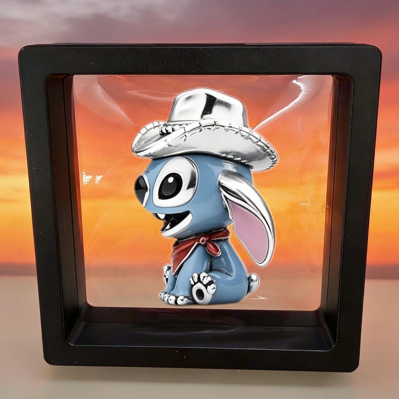 May include: A blue character figurine wearing a silver cowboy hat and a red neckerchief, suspended inside a square black floating display frame. The character has large black eyes, a wide open mouth, and pink-lined ears, presented in a clear case.
