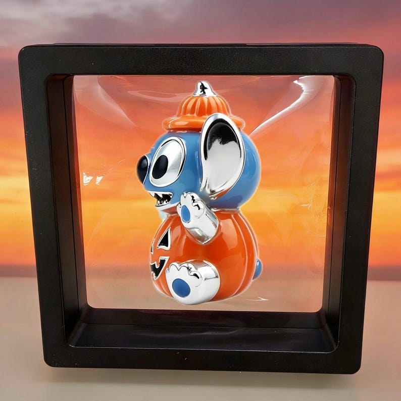 May include: A blue and orange character figurine wearing a pumpkin hat and body, displayed inside a square black floating frame. The character has large silver ears, a silver face, and a carved jack-o'-lantern design on its orange torso.