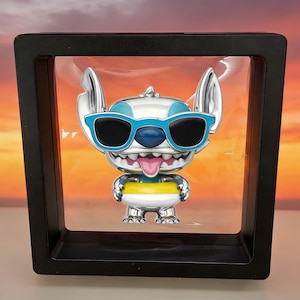 K&ouml;nnte beinhalten: Eine silberfarbene Sammlerfigur der Figur Stitch mit gro&szlig;en Ohren, blauer Sonnenbrille und herausgestreckter rosa Zunge, die in einem quadratischen schwarzen Schwebekader pr&auml;sentiert wird. Die Figur tr&auml;gt einen gelb-wei&szlig;en Schwimmring um die Taille.
