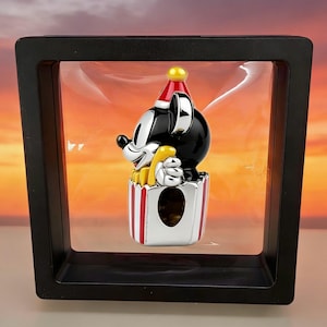 Op de afbeelding: Een zwart-wit Mickey Mouse-beeldje met een rode en gele feesthoed, met een gele popcorncontainer. Het beeldje wordt weergegeven in een zwarte schaduwdoos met een heldere voorkant.