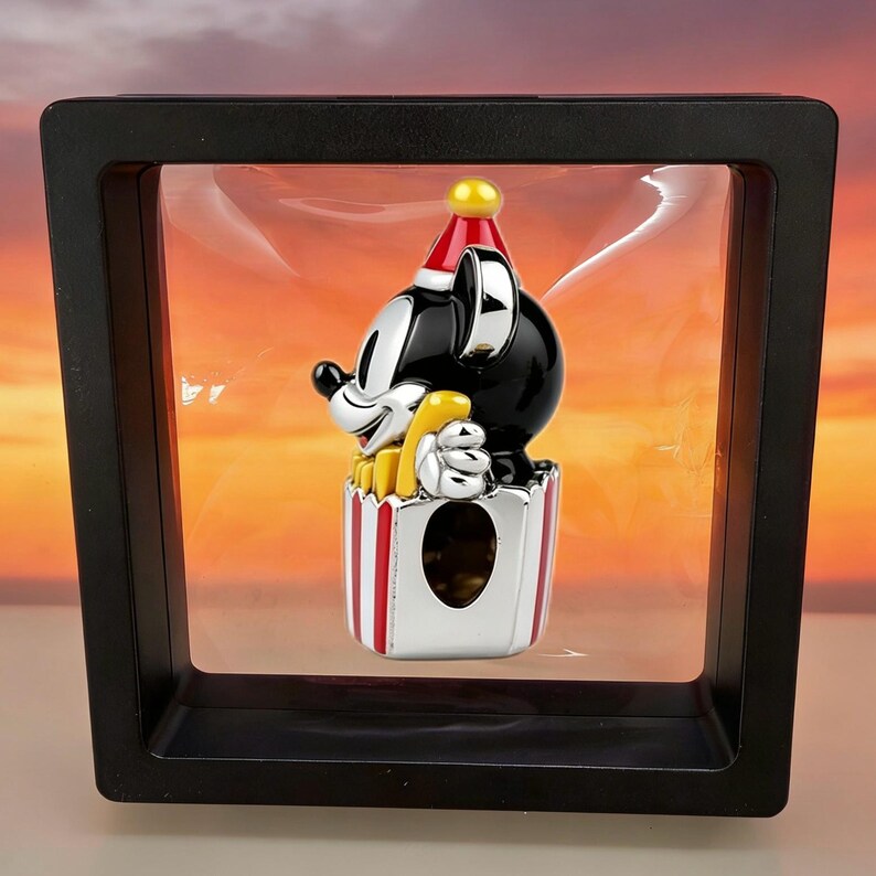 Op de afbeelding: Een zwart-wit Mickey Mouse-beeldje met een rode en gele feesthoed, met een gele popcorncontainer. Het beeldje wordt weergegeven in een zwarte schaduwdoos met een heldere voorkant.