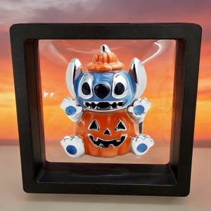 May include: A blue character figurine wearing an orange pumpkin hat and sitting inside an orange pumpkin body with a carved jack-o'-lantern face. The figurine is suspended within a square black floating display frame with clear plastic film.