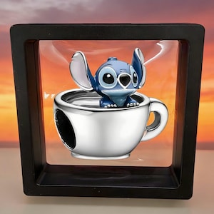 K&ouml;nnte beinhalten: Ein silberfarbener Charm in Form einer Teetasse mit einer blau-wei&szlig;en Figur mit gro&szlig;en Ohren und schwarzen Augen, die darin sitzt. Der Charm ist in einem quadratischen schwarzen Rahmen mit einer transparenten, flexiblen Membran befestigt.