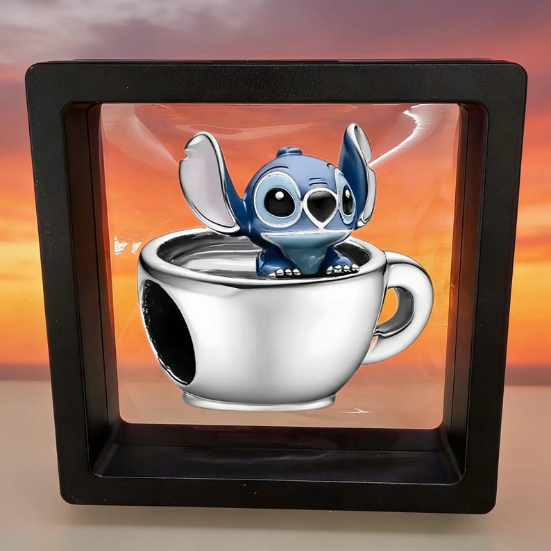 K&ouml;nnte beinhalten: Ein silberfarbener Charm in Form einer Teetasse mit einer blau-wei&szlig;en Figur mit gro&szlig;en Ohren und schwarzen Augen, die darin sitzt. Der Charm ist in einem quadratischen schwarzen Rahmen mit einer transparenten, flexiblen Membran befestigt.