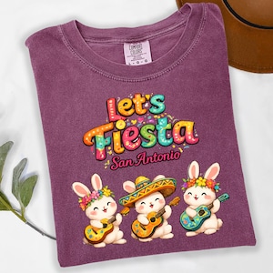 May include: A purple t-shirt with the words "Let's Fiesta San Antonio" in colorful lettering. The shirt features three cartoon bunnies playing guitars and wearing flower crowns and sombreros. The shirt is a Comfort Colors brand.