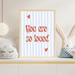 Puede incluir: Un estampado enmarcado con las palabras "You are so loved" en rojo sobre un fondo de rayas azules y blancas. Dos corazones rojos acentúan el texto. El estampado está en un marco de madera clara.