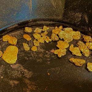 Puede incluir: Un conjunto de pepitas de oro en una sartén negra. Las pepitas varían en tamaño y forma, mostrando un color dorado intenso. La sartén tiene un diseño curvo, con una superficie oscura y texturizada. Las pepitas están dispersas.