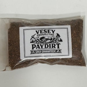 Puede incluir: Una bolsa de plástico transparente llena de tierra de pago marrón. Una etiqueta blanca con texto negro dice "VESEY PROSPECTING PAYDIRT GOLD GUARANTEED!" con un gráfico de un pico, una sartén y una pala. La bolsa está sellada.