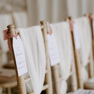 May include: A row of wooden chairs decorated for a wedding ceremony. Each chair has a white fabric draped over the back and a tag that reads "Reserved for the Mother of the Groom". A pink ribbon is tied to the top of each chair.