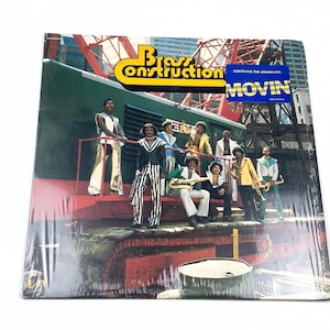 May include: A vinyl record cover for Brass Construction featuring a group of musicians standing on a green industrial train car. The text reads Brass Construction and contains the smash hit Movin. The background shows red construction cranes.