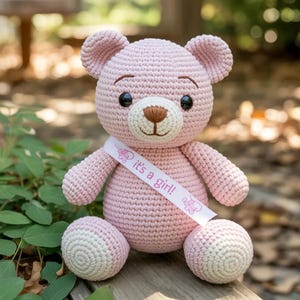 May include: A handmade pink crochet teddy bear with a white ribbon that reads "it's a girl!". The bear has black eyes, a brown nose, and white accents on its paws. The bear is sitting on a wooden surface.