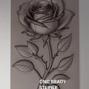 May include: A detailed, monochromatic relief carving of a rose in full bloom, complete with leaves and thorns, set against a rectangular background. The text "CNC READY STL FILE VETRIC & VCARVE" is visible at the bottom.