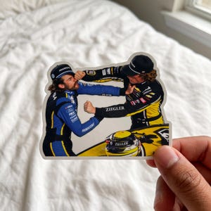 May include: A die-cut sticker depicting two race car drivers in professional racing suits engaged in a physical altercation. One driver wears a blue and black suit, while the other wears a black and yellow suit with the text ZIEGLER visible.