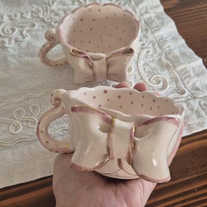 Puede incluir: Dos tazas de té de cerámica rosa con diseños en forma de lazo y detalles dorados. Las tazas tienen un estampado de lunares en el interior y un asa curvada. Las tazas de té están sobre un mantel de encaje blanco.